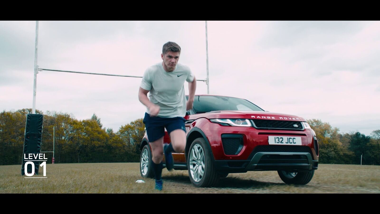Rugby player Owen Farrell standing beside a red Range Rover Evoque on a rugby pitch with goalposts