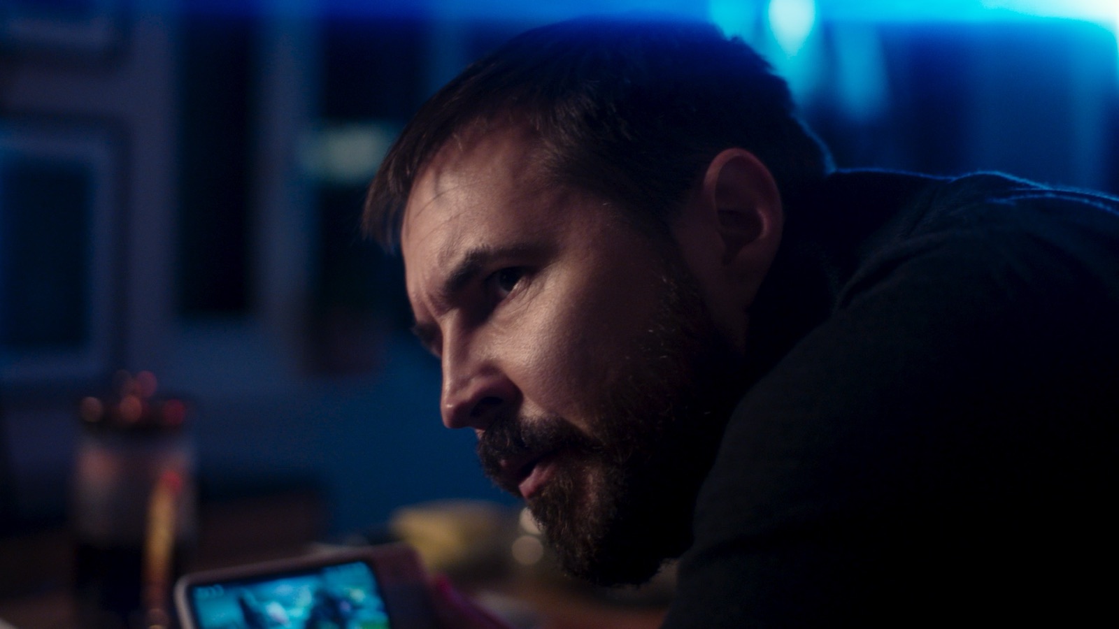 Martin Compston leaning forward studying phone screen in cool blue lighting