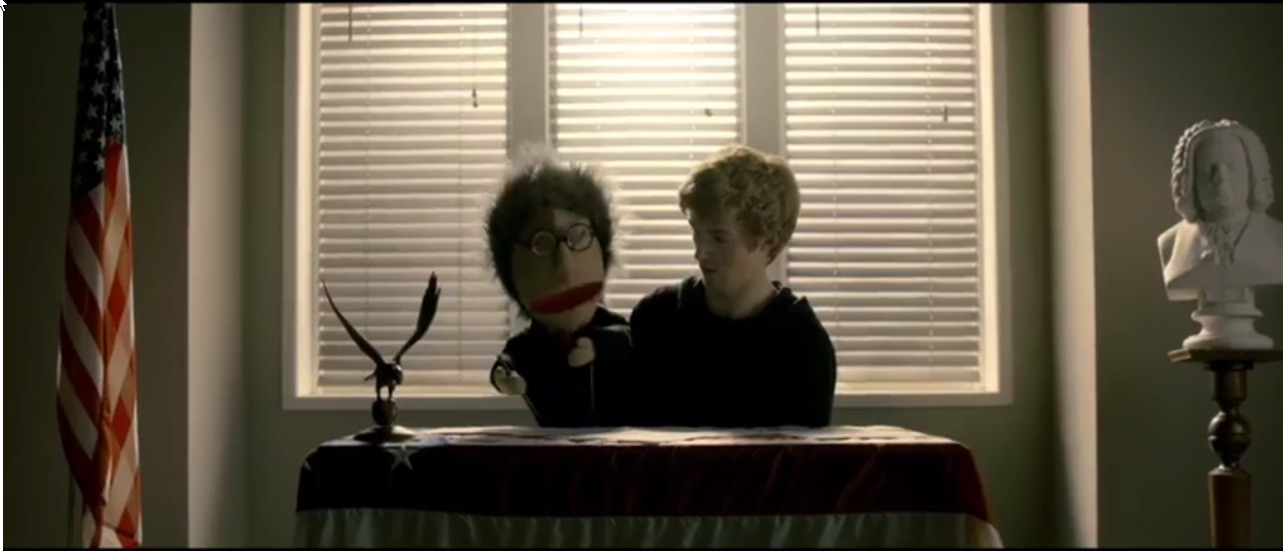 Two puppets seated at a desk with an American flag and bust in a muted desaturated room
